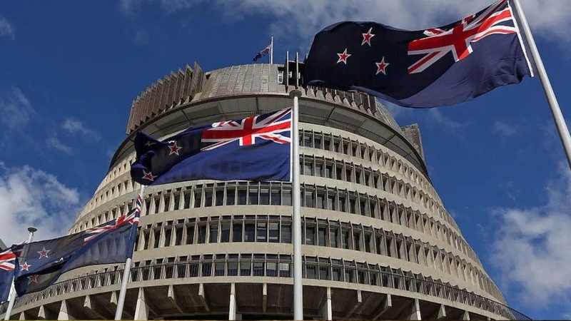 New Zealand flags fly on tall poles in front of the Beehive, the circular parliament building in Wellington, under a bright blue sky